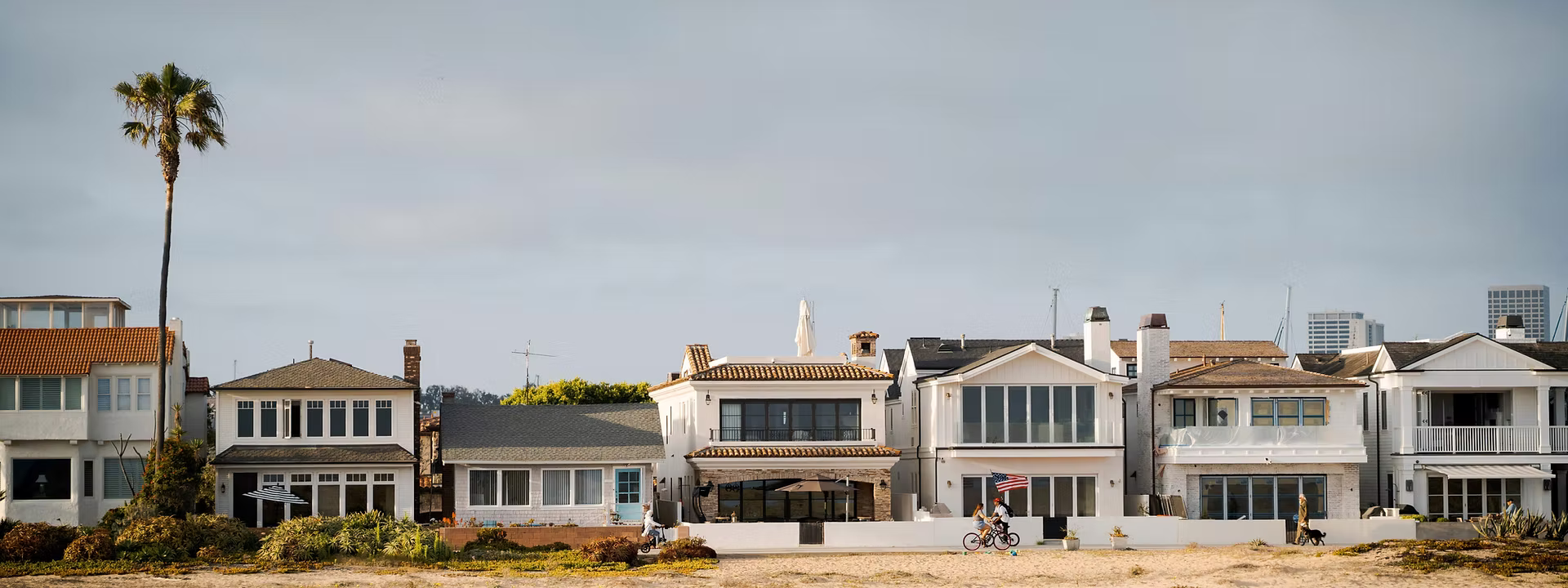 Beachfront houses with balconies, a lone palm tree, two people biking, and a person walking a dog. Overcast sky sets a calm mood.