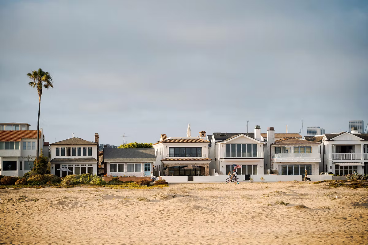 A row of luxury modern and Mediterranean-style multi-story homes lining the Hermosa Beach Strand, with the paved pedestrian path in the foreground and a sandy beach under a clear blue sky.