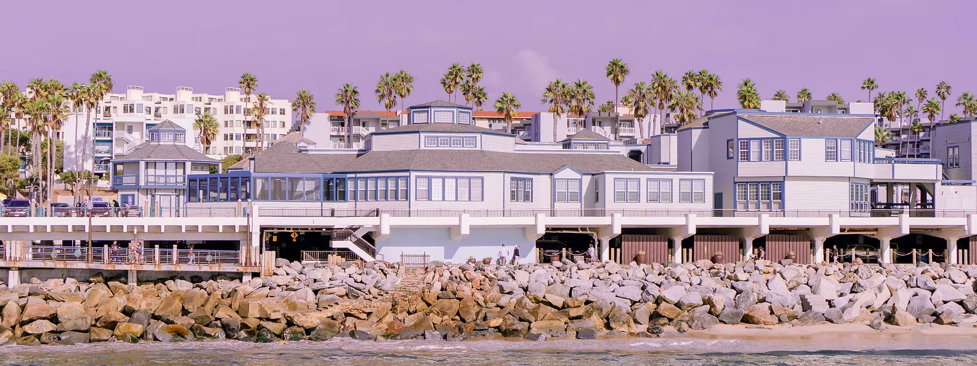 Coastal scene showing beachfront buildings with gray roofs, rocky shore, palm trees, and a calm blue sea under a soft purple sky.